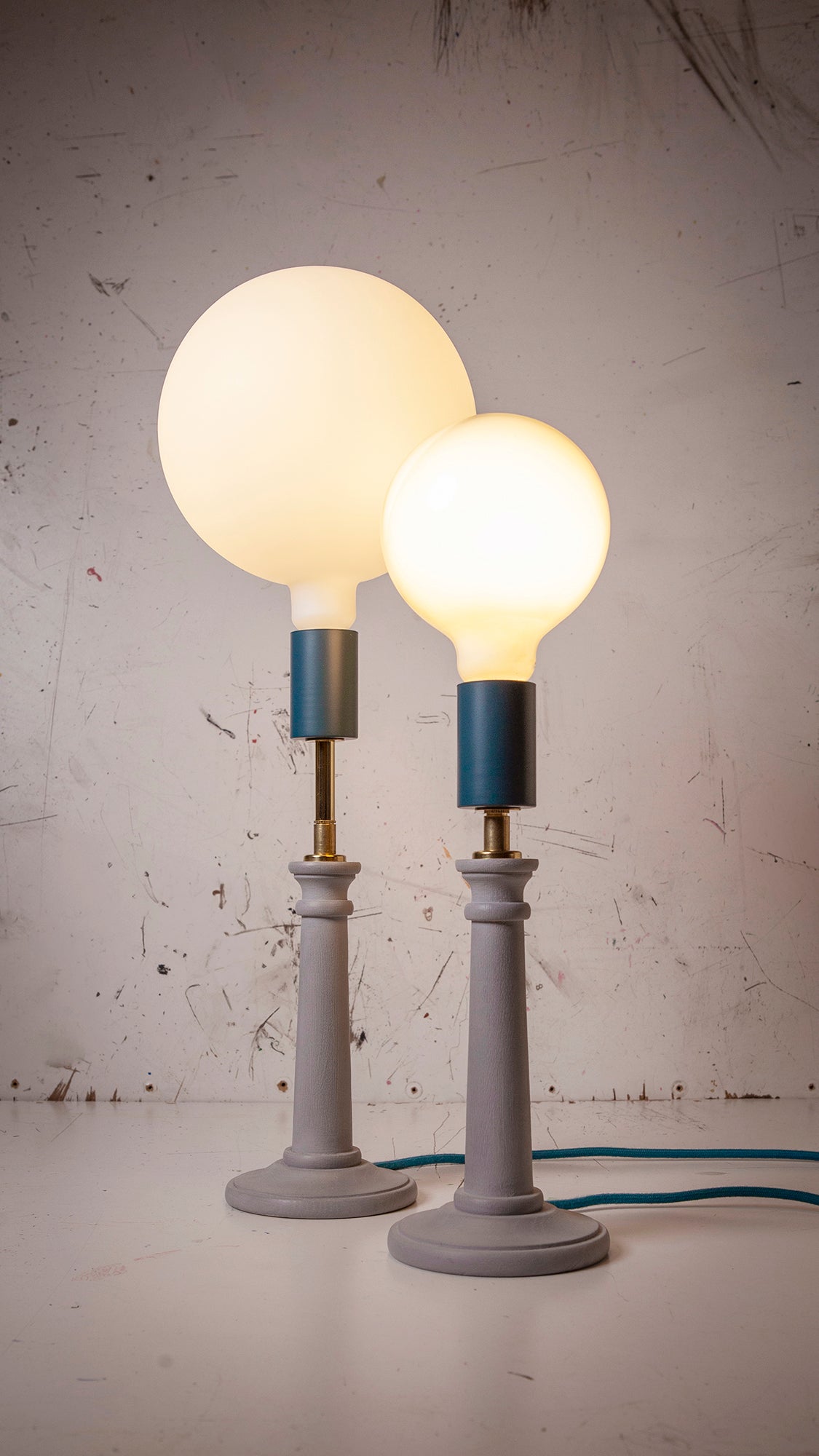 Two table lamps with round light bulbs on light blue stands against a textured industrial white wall.